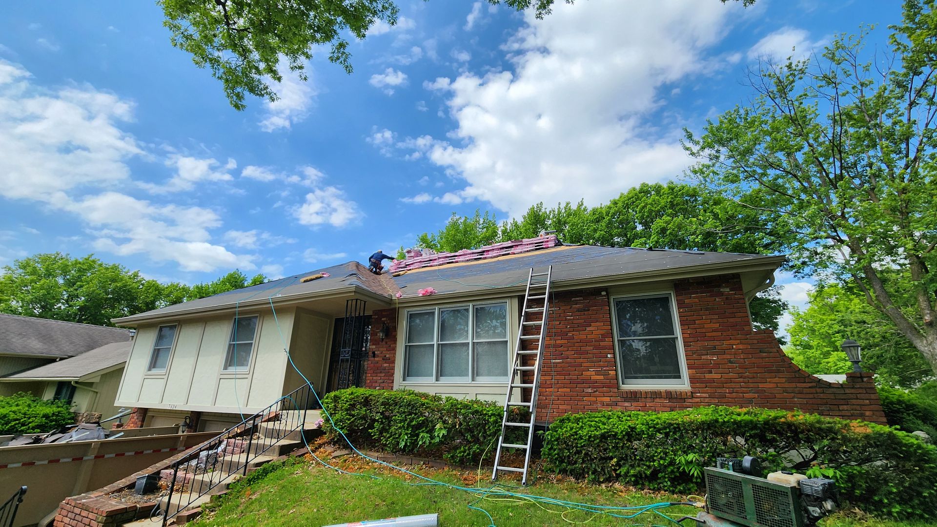 House with a partially replaced roof; ladder on the side. Blue sky, green bushes, and a sunny day.