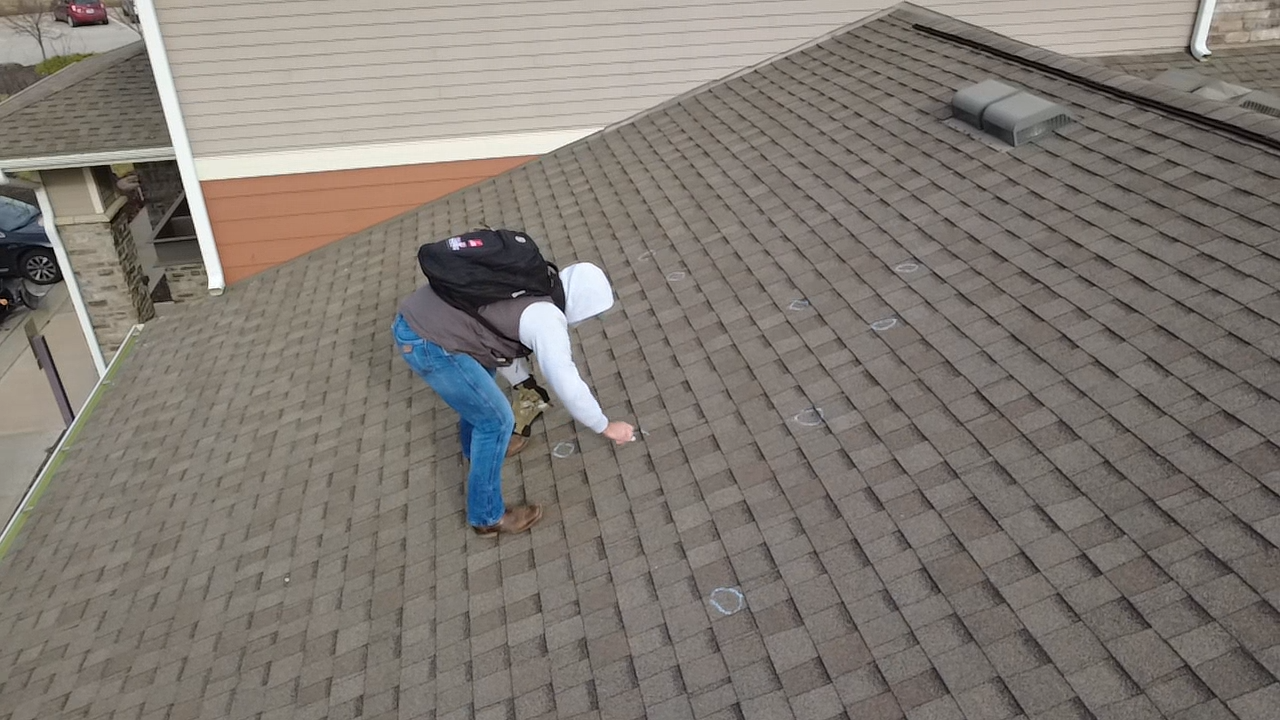 Person with backpack on a brown shingled roof, reaching towards shingles. Beige exterior, overcast sky.