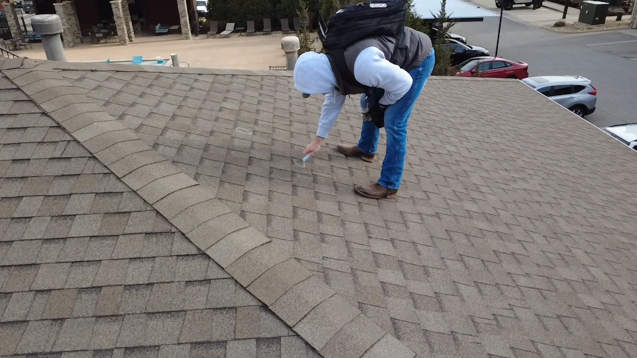 Person on a brown shingled roof inspecting something. They wear a backpack, jeans, and a white hat.