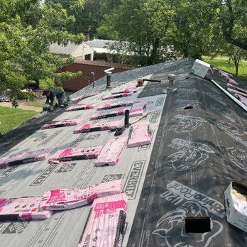 A worker installs shingles on a sloped roof covered with underlayment and bundles of roofing materials.