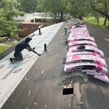 A roofer installs underlayment on one side of a pitched roof, while bundles of pink shingles are lined up on the other side.