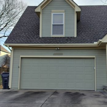 A sage green house exterior featuring a two-car garage with a matching dormer window above and a dark gray driveway.