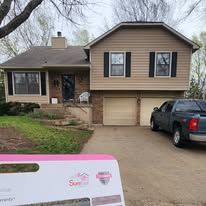 A two-story house with tan siding and a brick lower level, featuring a driveway with a parked dark pickup truck.