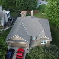 Aerial view of a residential house with a gray shingled roof, a front garage, and two parked cars in the driveway.