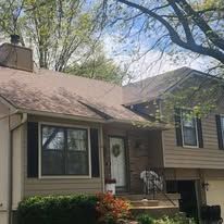 A two-story house with tan siding, a brown roof, and a chimney, viewed from the front yard under a tree.
