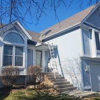 A blue two-story house with a ladder leaning against the siding near the front entrance steps under a clear blue sky.