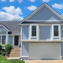 A two-story light blue house with a white garage door, a prominent front gable, and a small porch with concrete stairs.
