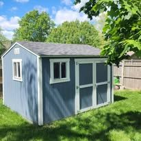 A blue storage shed with white trim and windows sits on a grassy lawn under a partially cloudy blue sky.