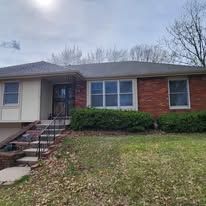 A single-story brick house with a brown shingled roof, a front porch with stairs, and a lawn covered in fallen leaves.