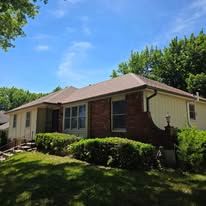 A single-story house with a brick and light-colored siding exterior, surrounded by green landscaping under a blue sky.
