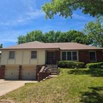 A split-level suburban house with a tan exterior, brick accents, a two-car garage, and a lush green lawn under a blue sky.