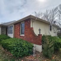 A one-story house with a brick facade on the left and light-colored vertical siding on the right, surrounded by bushes.