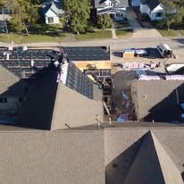 An aerial view shows workers on a multi-sloped residential roof during construction, with materials visible on the ground.