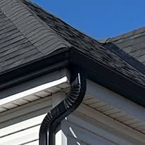 A close-up view of black metal rain gutters and a matching downspout attached to the corner of a house with gray shingles.