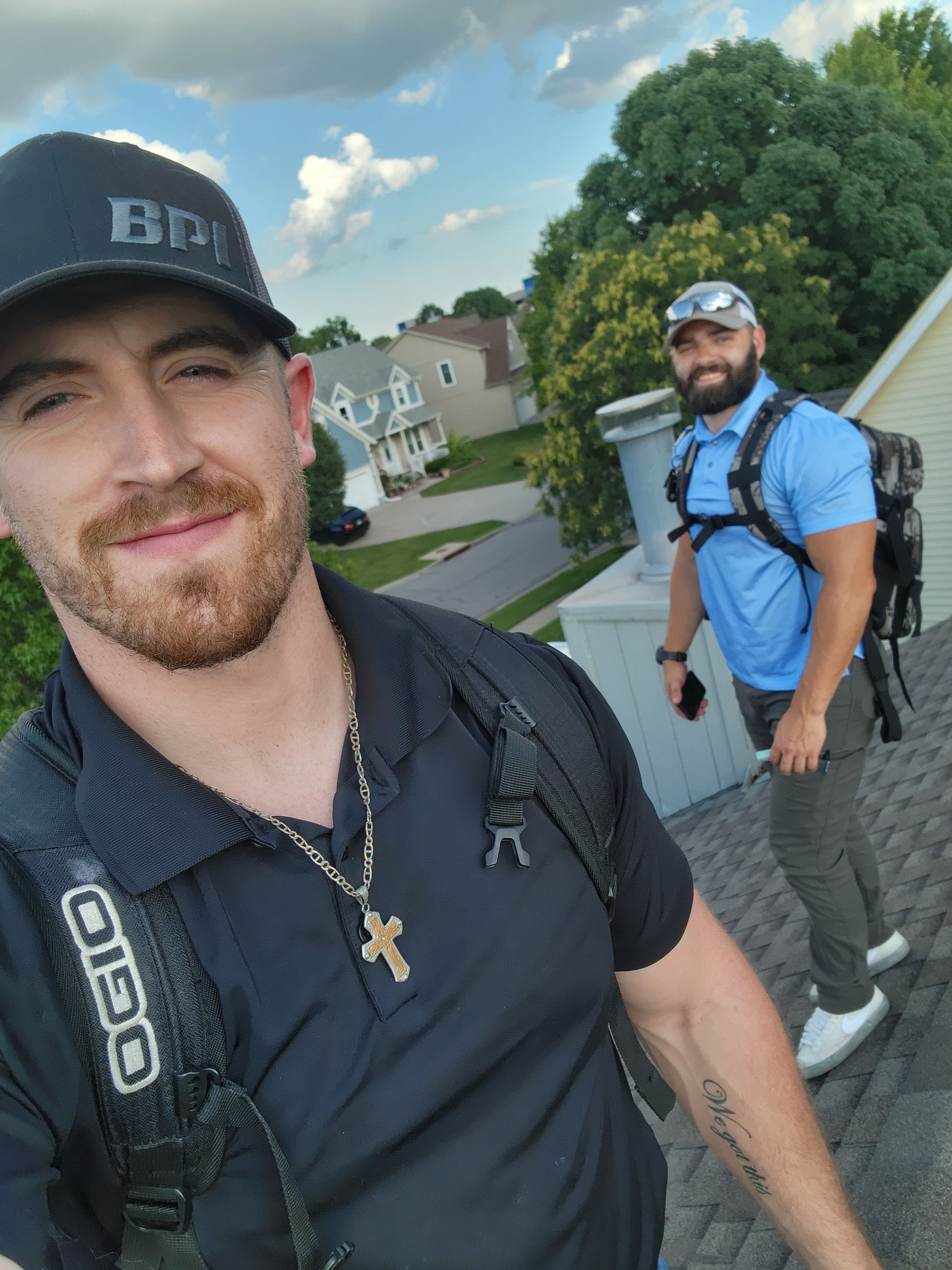 Two men on a roof; one takes a selfie, the other wears a backpack. Blue sky, suburban setting.