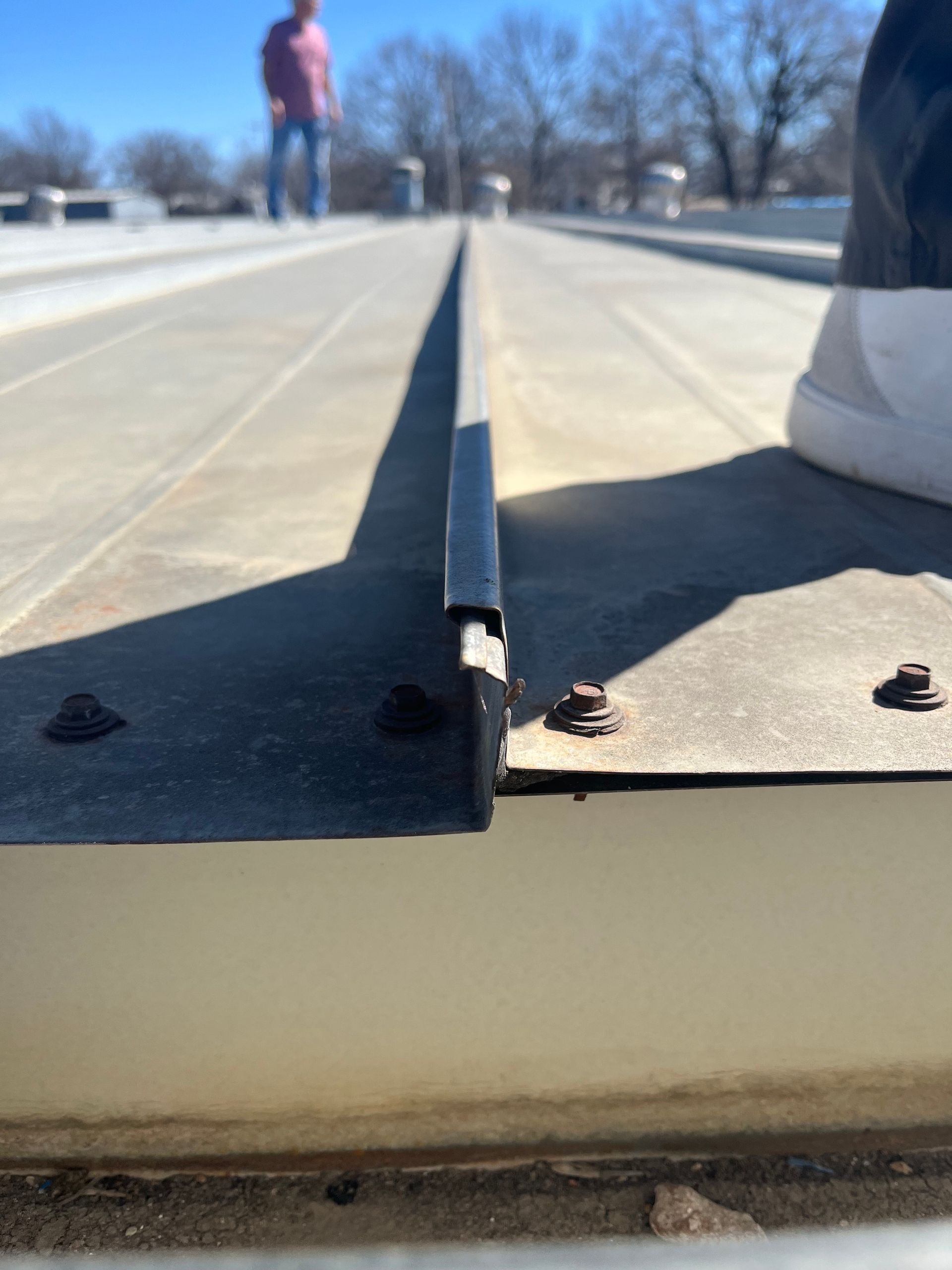 Close-up of a metal roof seam with visible bolts. A person is standing in the distance.