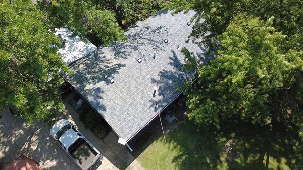 A high-angle aerial view of a single-story house with a gray shingled roof, surrounded by trees, with a truck parked below.
