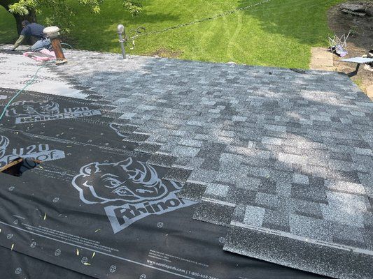 A worker installs gray asphalt shingles over black roofing underlayment on a residential roof.