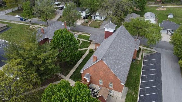 An aerial view of a red brick church building with a gray roof and steeple, surrounded by trees, a sidewalk, and a parking lot.