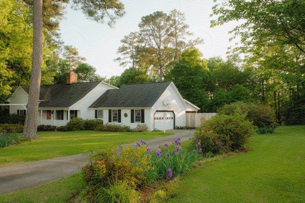 A white house with a dark shingled roof, set in a lush green lawn surrounded by mature trees.
