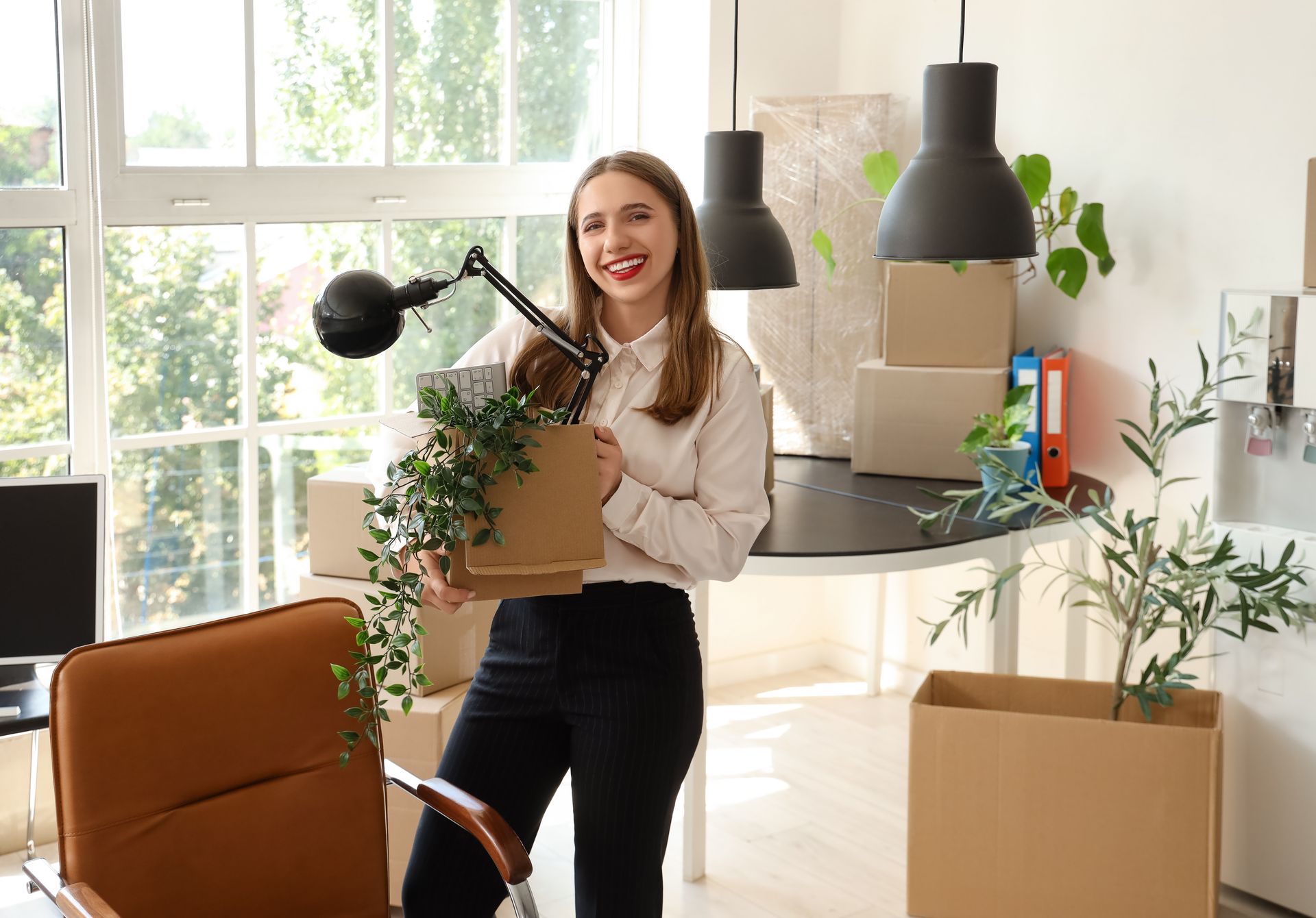 Smiling woman carrying a box in a bright room with houseplants and pendant lights