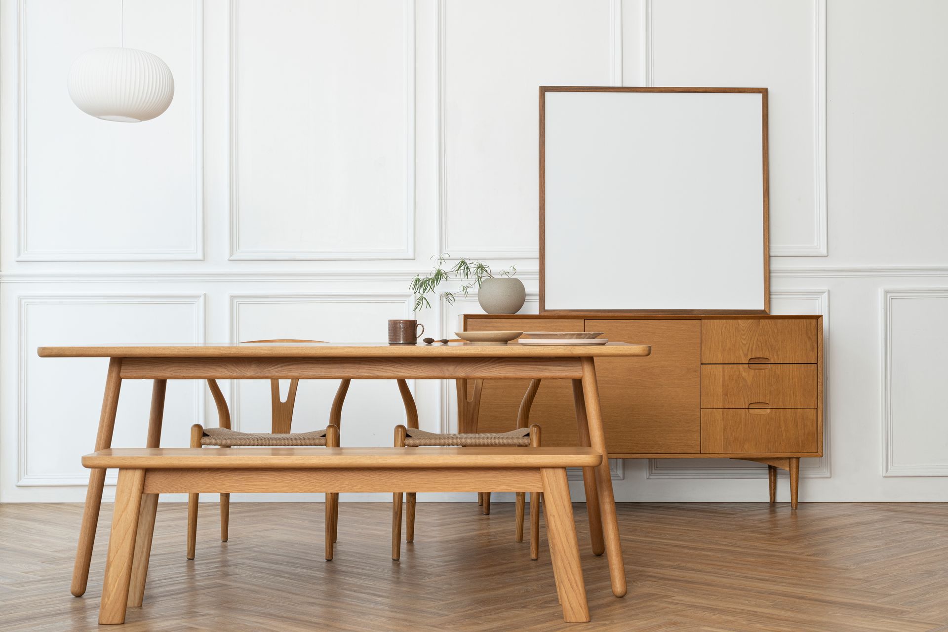 Minimalist dining room with wooden table, chairs, sideboard, and framed mirror on a white paneled wall