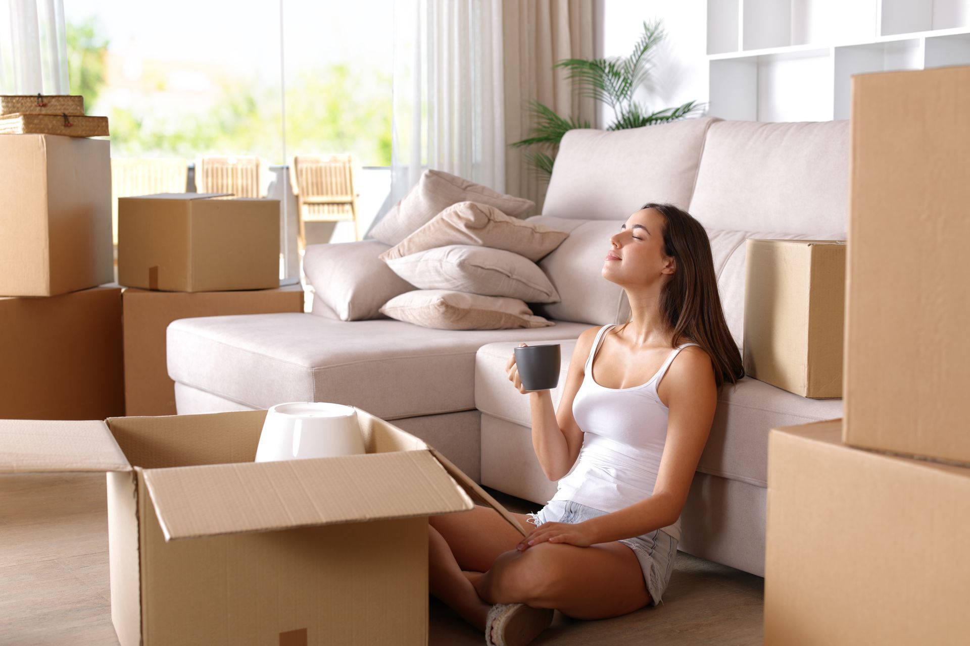 Woman sitting on the floor among moving boxes beside a sofa in a bright living room