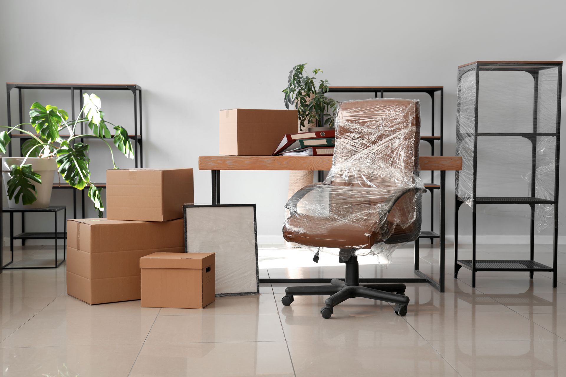 Packed moving boxes and wrapped office chair beside a desk in a bright room