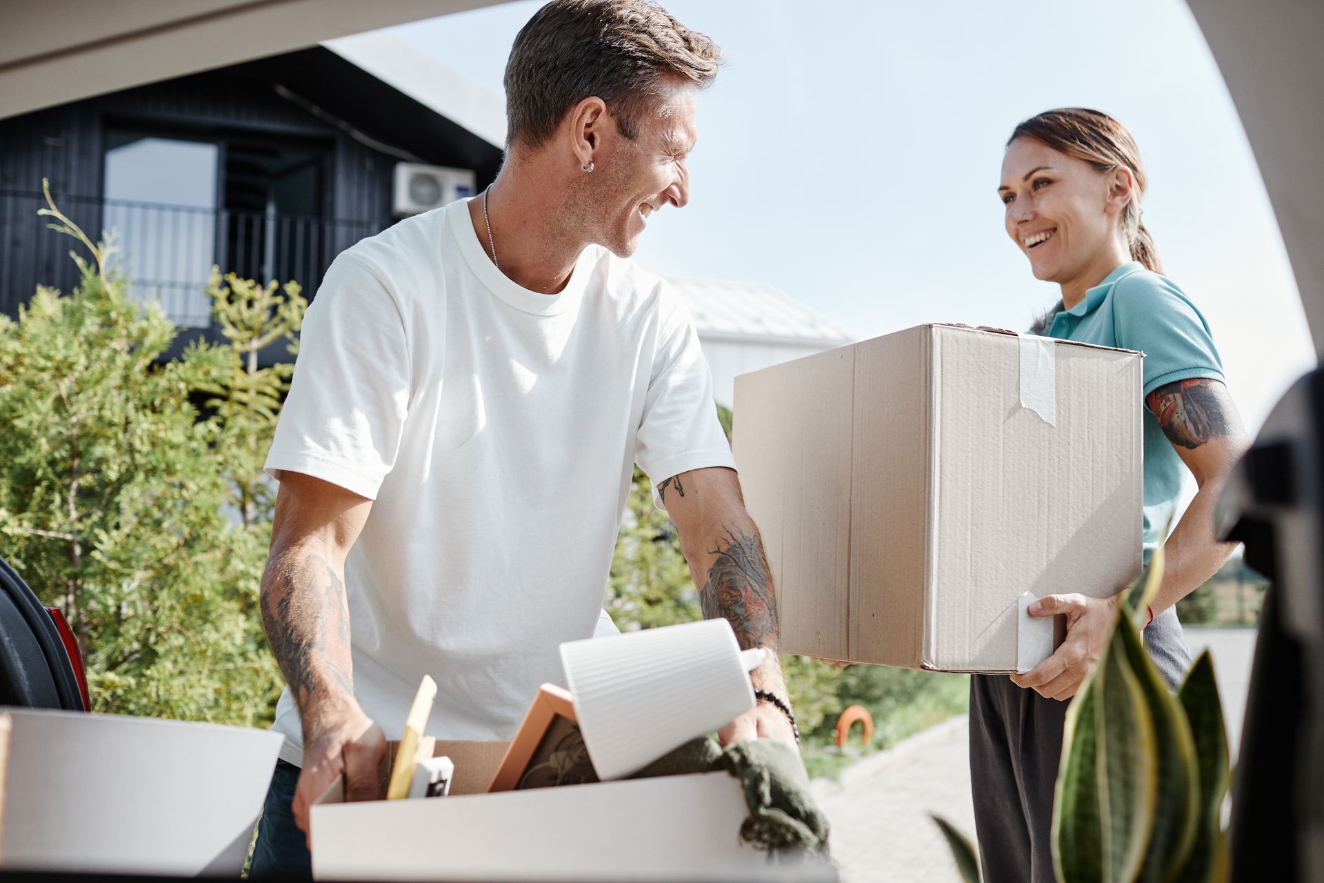 Two people carrying moving boxes outside a house, smiling in a sunny yard