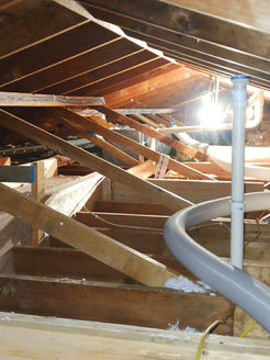 A view inside an attic with wooden rafters, insulation, a vertical white vent pipe, and a grey flexible hose.