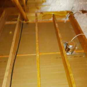 An attic crawl space showing wooden joists, brown insulation boards, wiring, and a recessed lighting fixture.