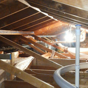 A view inside an attic showing wooden roof rafters and framing with a vertical white pipe standing in the foreground.
