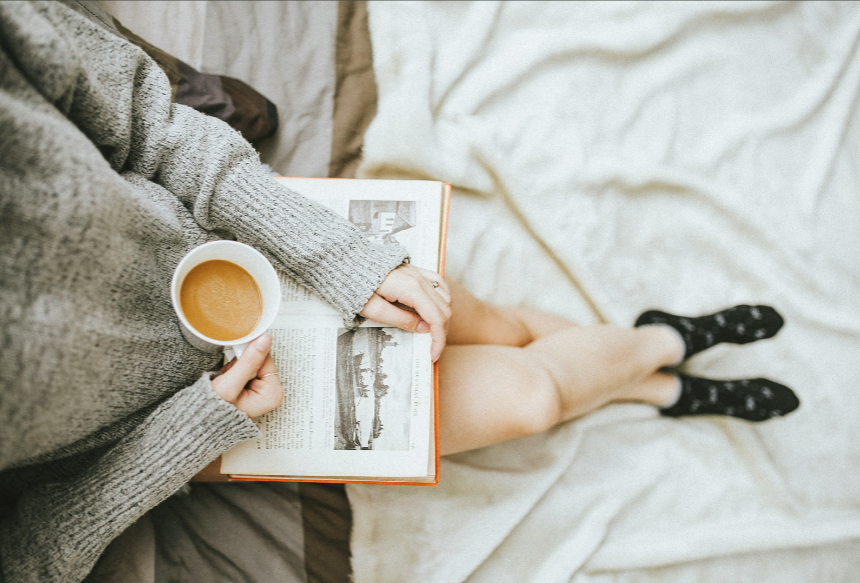 A woman is sitting on a bed reading a book and holding a cup of coffee