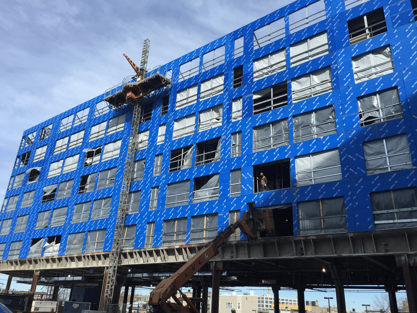 Blue-wrapped building under construction with scaffolding and a crane lift on the facade