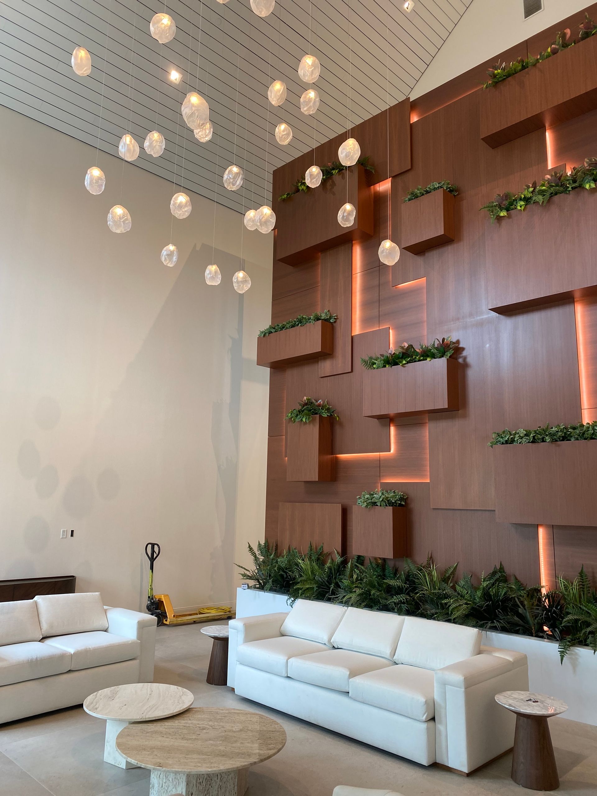 Modern lobby with white sofas, round tables, hanging lights, and a wood wall with green plants.