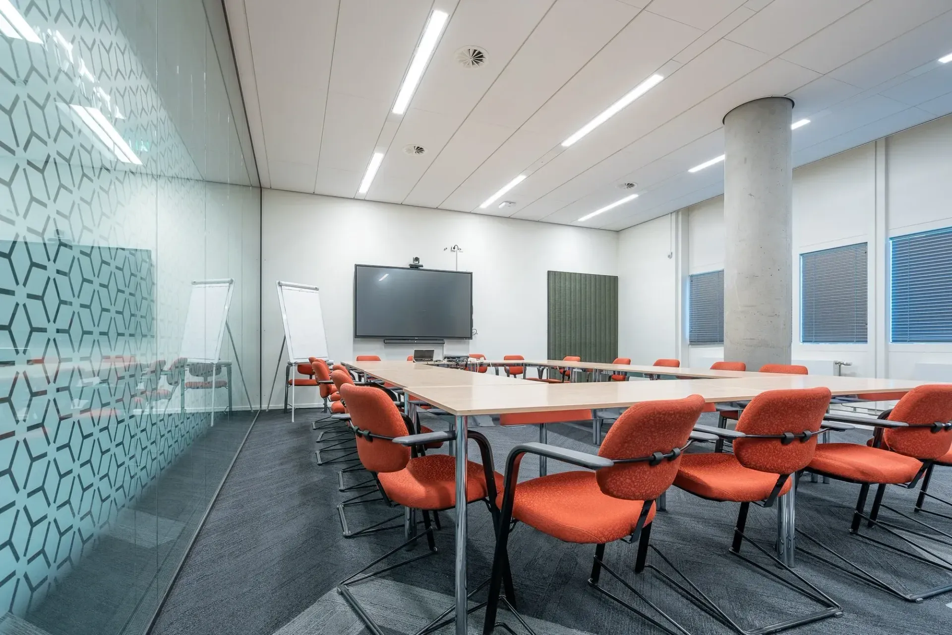 Empty modern conference room with orange chairs, long table, wall-mounted screen, and patterned glass walls