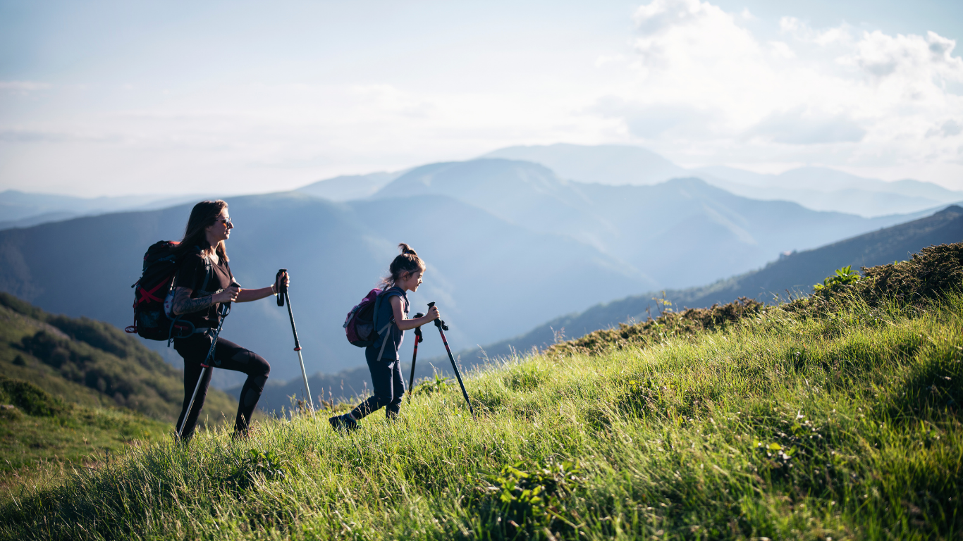Two women with hiking packs and walking sticks walk in the grassy meadow on a mountain-side