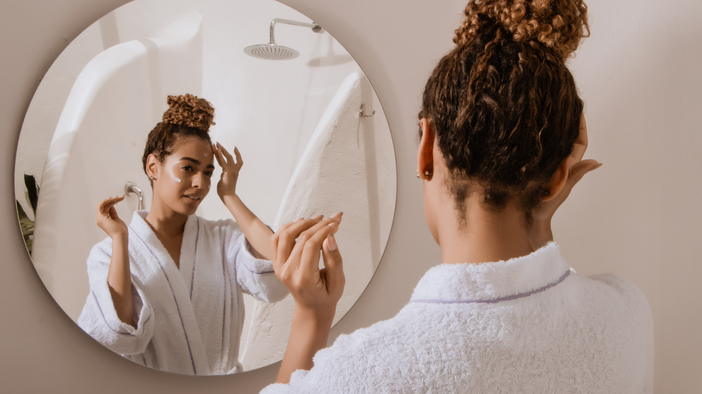 A woman in a white robe touches her face and looks at herself in a circular, wall hanging mirror