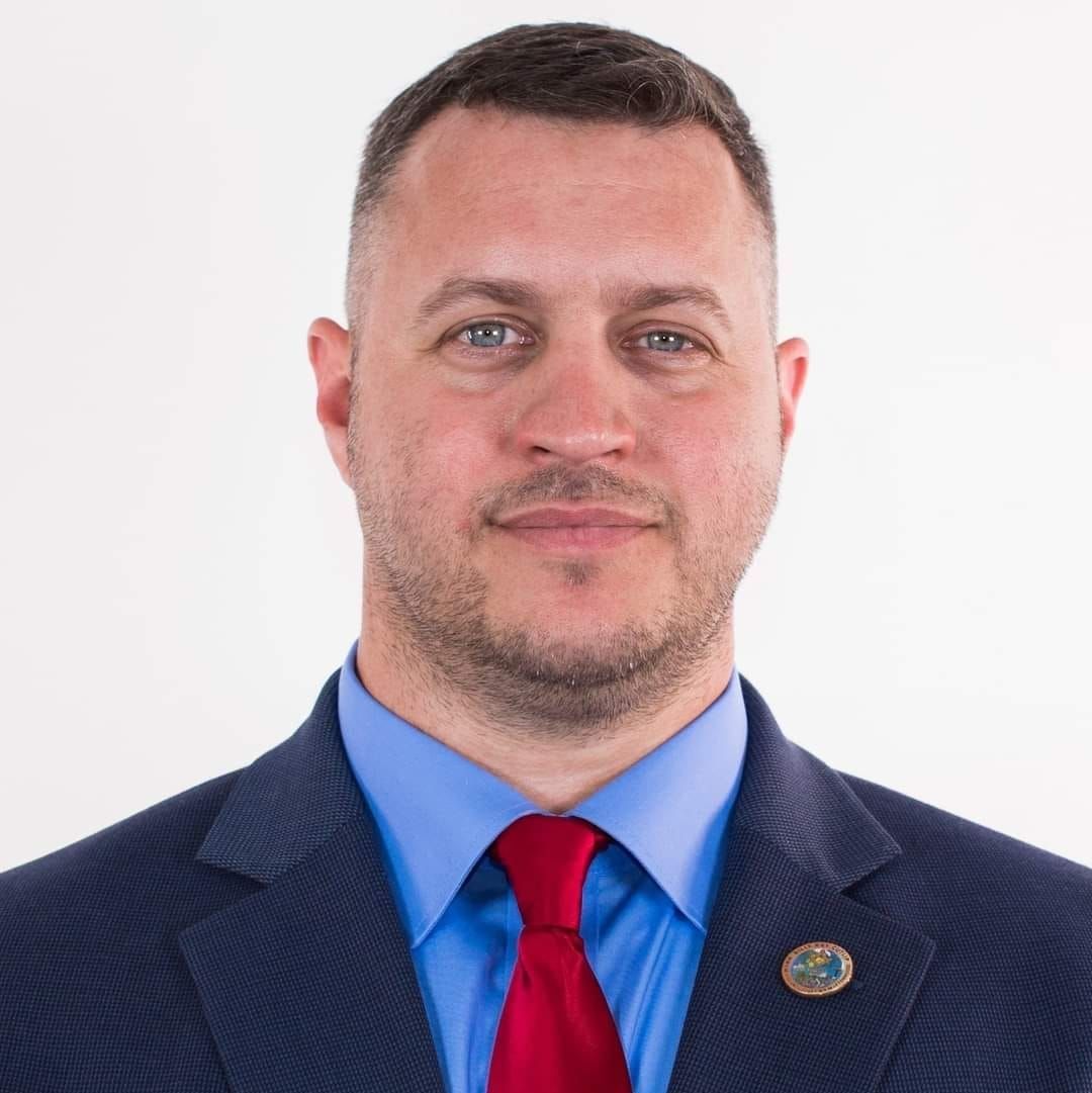 Studio headshot of a man in a blue shirt, red tie, and dark suit jacket on a white background