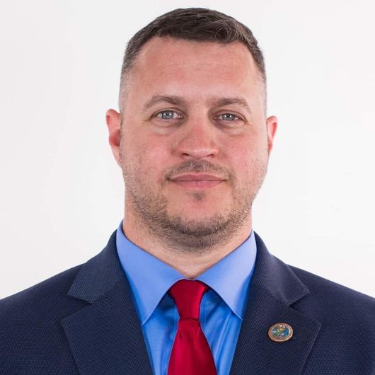 Formal head-and-shoulders portrait of a man in a blue shirt, red tie, and navy suit jacket on a white background