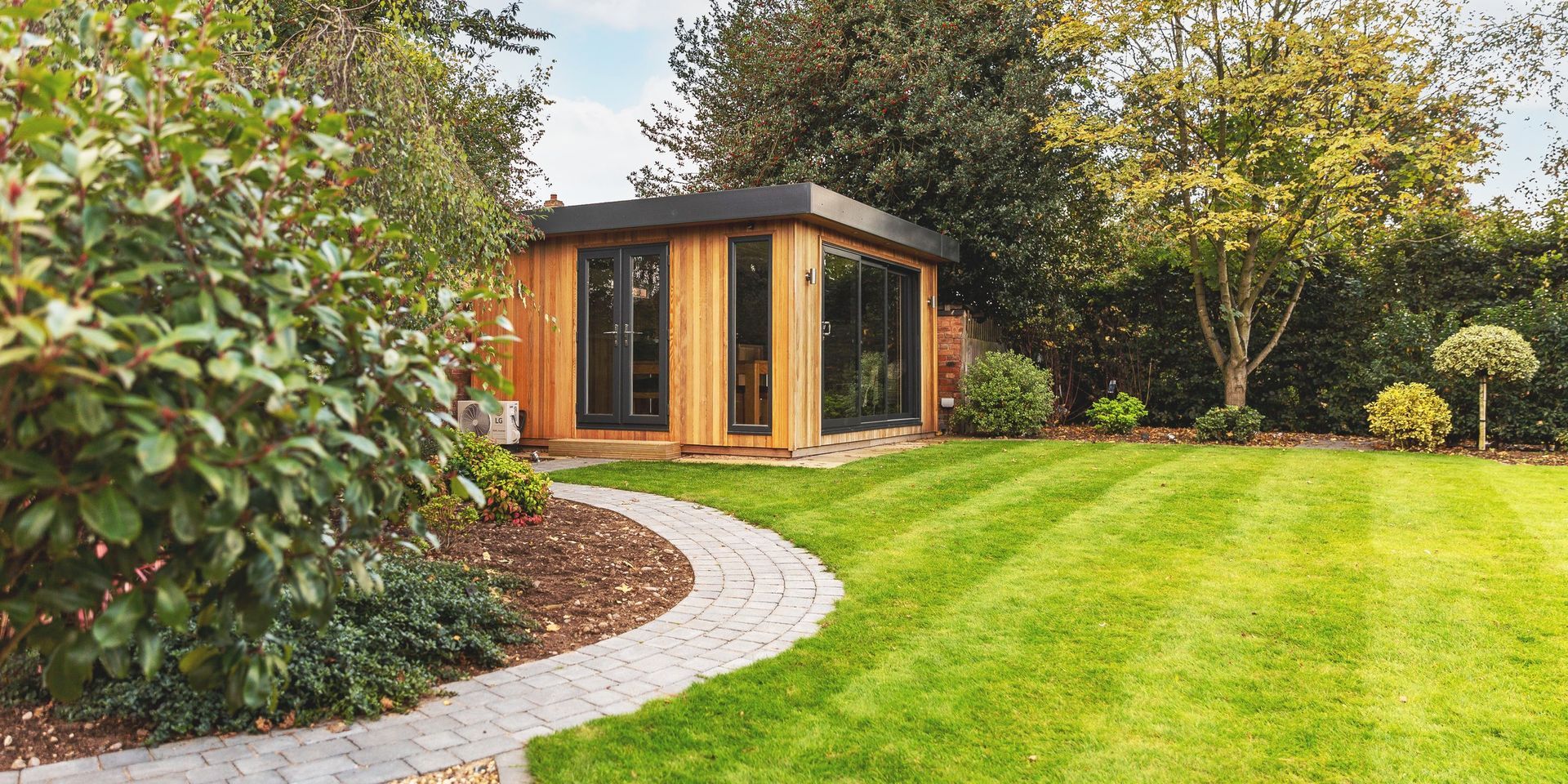 A wooden garden office sits in a green lawn. A stone path leads to the structure. Lush greenery surrounds the yard.