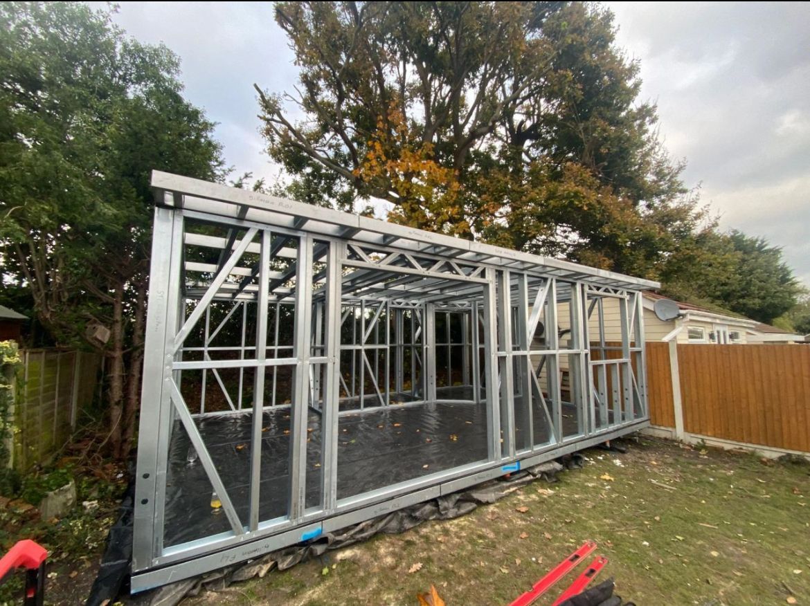 Metal frame of a building under construction, set in a backyard. Trees and a fence are visible in the background.