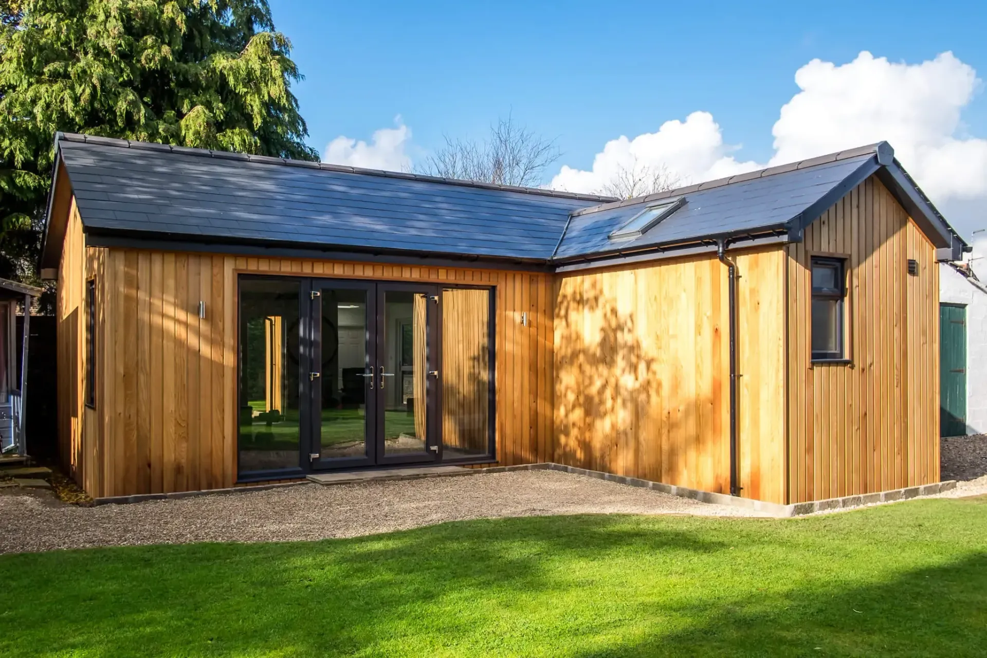 Wooden garden studio with a slate roof, glass doors, and small window on a grassy lawn.