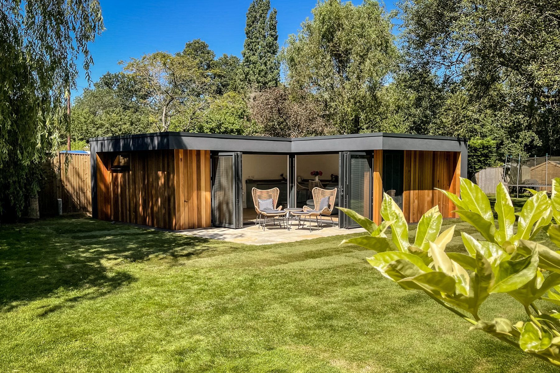Wooden garden structure with open front, two chairs inside, surrounded by trees and grass.