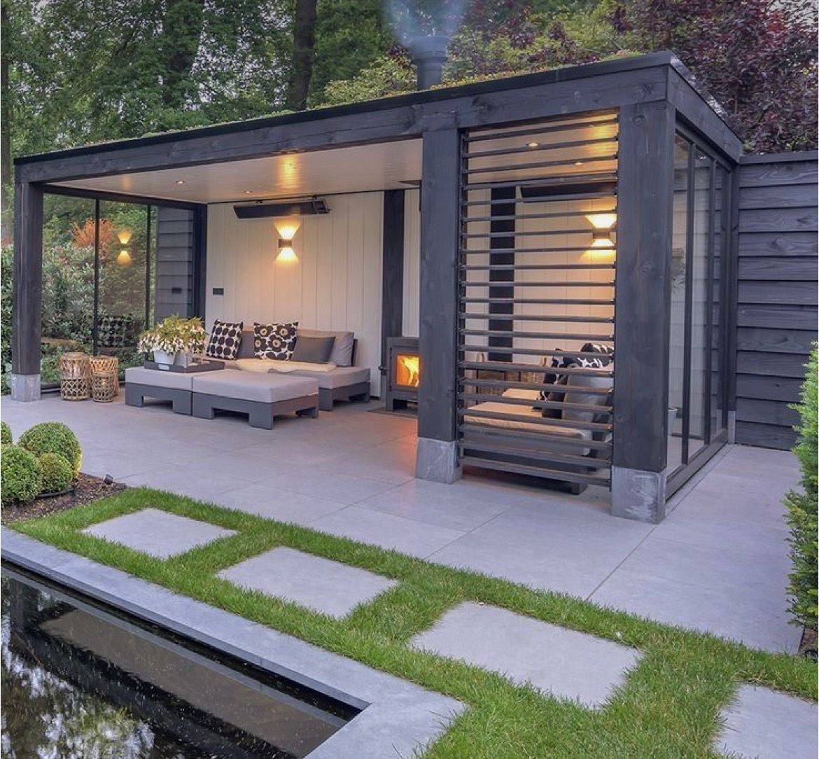 Outdoor patio with a seating area, fireplace, and stepping stones alongside a body of water.