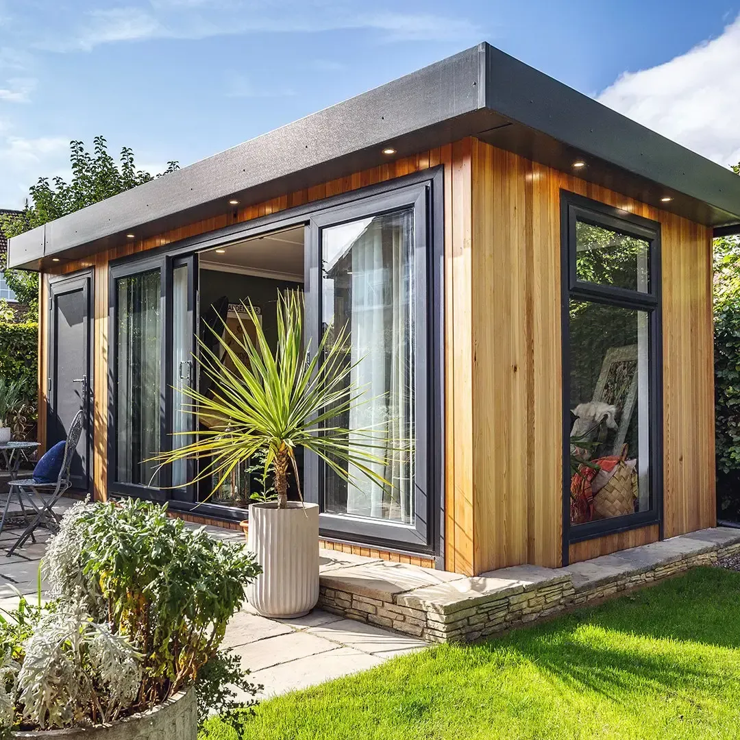 Wooden backyard studio with glass doors and a black roof, surrounded by plants and a patio.