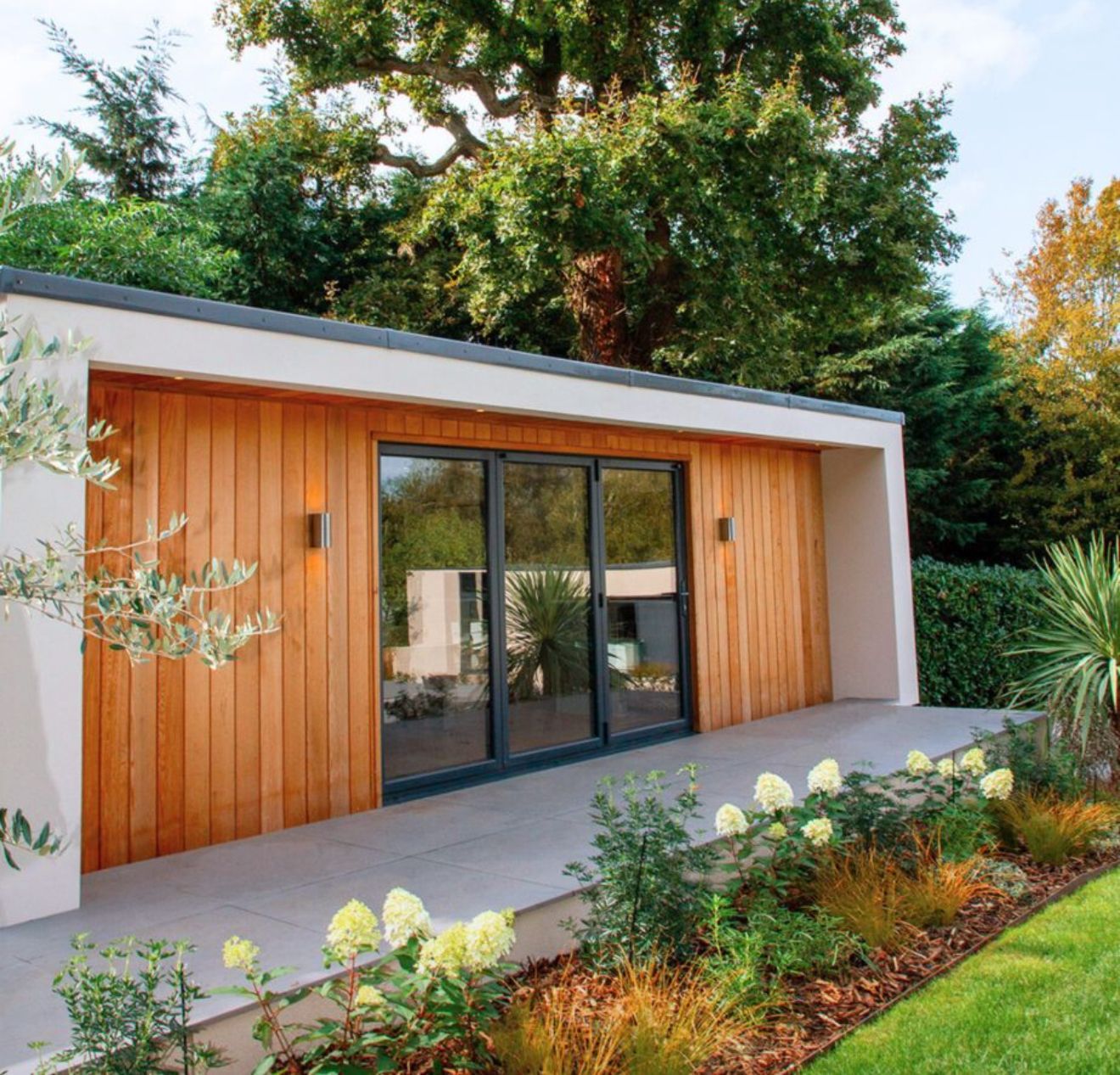 Modern garden building with wooden siding and glass doors, surrounded by landscaping.