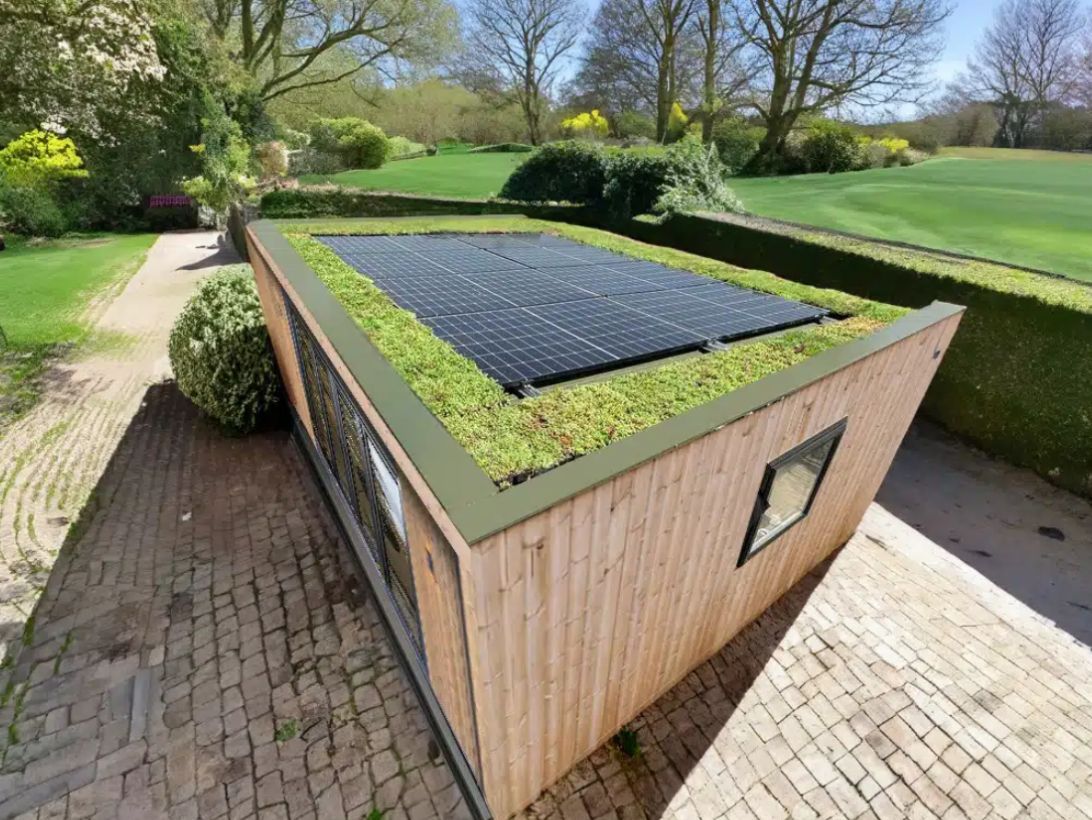 Green-roofed wooden shed with solar panels, set on a brick patio, surrounded by green grass and trees.