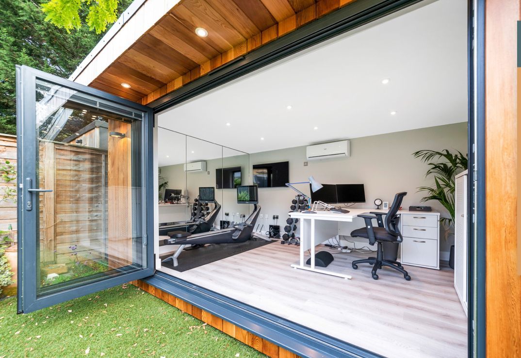 Modern home office with open glass doors, gym equipment, desk, and greenery.