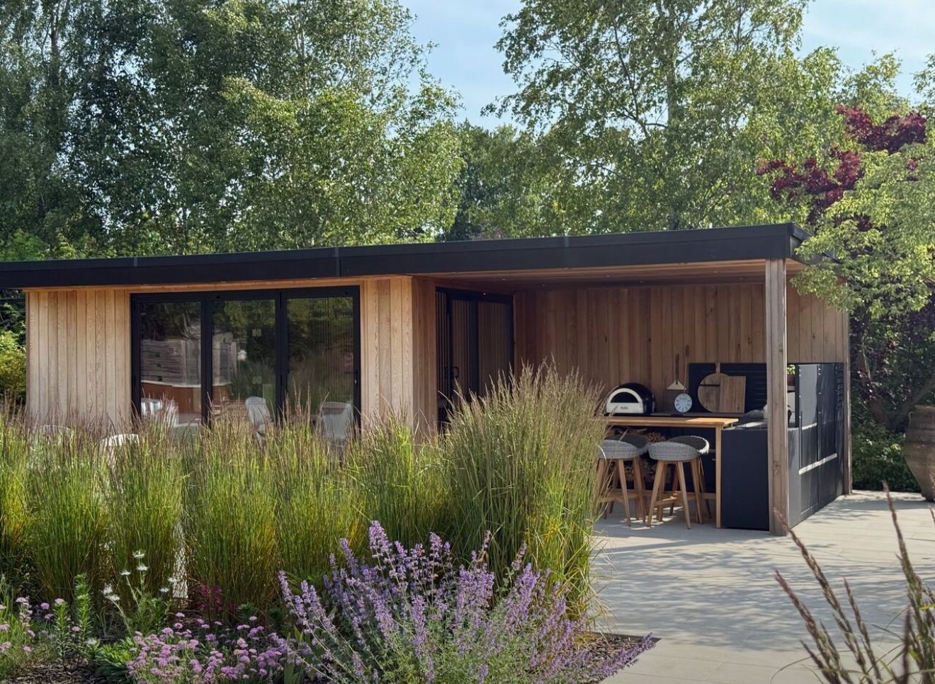 Wooden outdoor kitchen with pizza oven and bar stools, surrounded by lavender and trees.